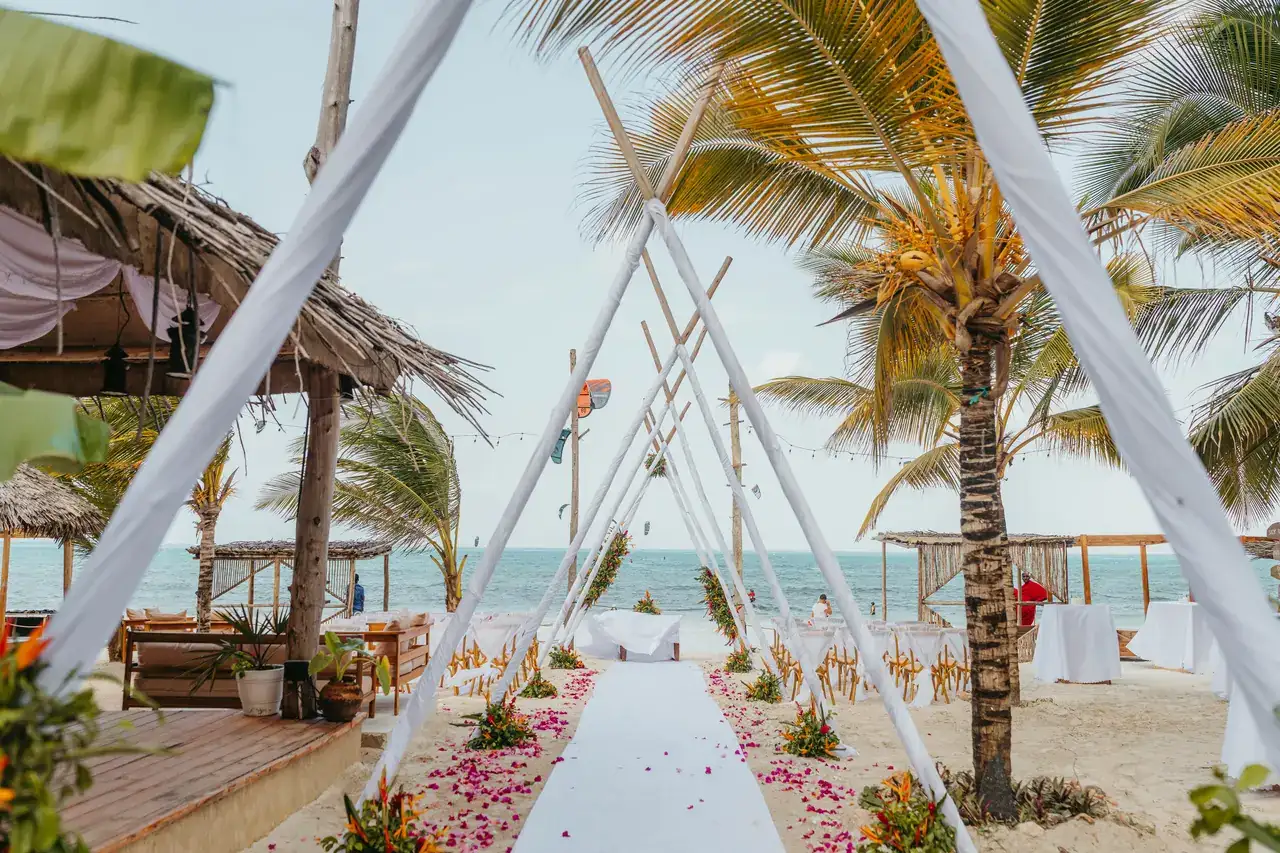 Draped white teepee ceremony arch on a beach, with a petal-lined aisle leading toward the ocean between palm trees